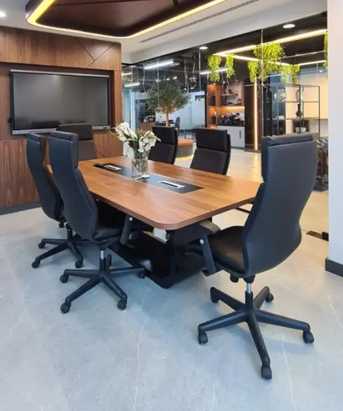 A modern conference room with a wooden table surrounded by six black office chairs. A flat screen TV is mounted on the wall, and there are decorative elements including a vase of flowers on the table and hanging plants near the windows.