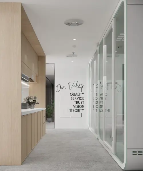 A modern office hallway with light wood paneling on the left and glass-paneled meeting rooms on the right. On the back wall, black text reads "Our Values: Quality, Service, Trust, Vision, Integrity." A small bouquet of flowers is on the countertop.