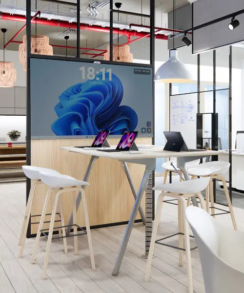 A modern office space featuring a high table with four white stools. Two laptops are on the table, displaying pink and blue screens. In the background, a large digital screen shows the time as 18:11. The room has contemporary decor and ample lighting.